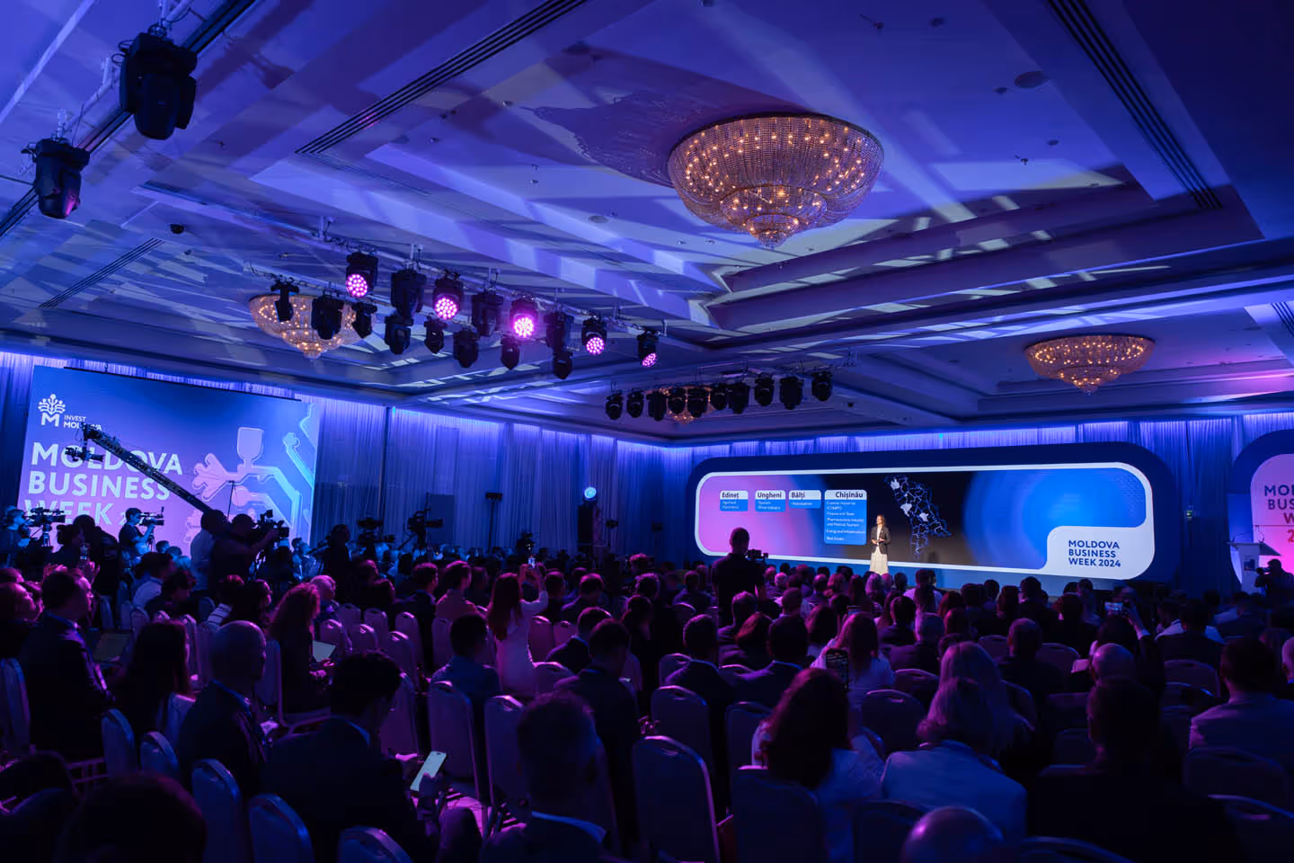 Конференц-зал с аудиторией на мероприятии Moldova Business Week 2024, человек выступает на сцене с большим экраном позади.