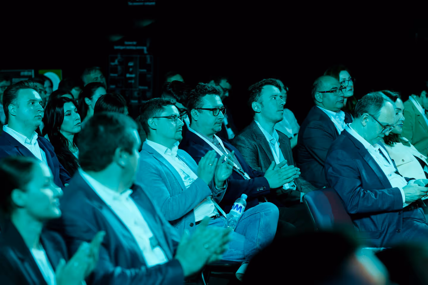 Группа взрослых, сидящих в рядах и аплодирующих во время мероприятия в помещении.