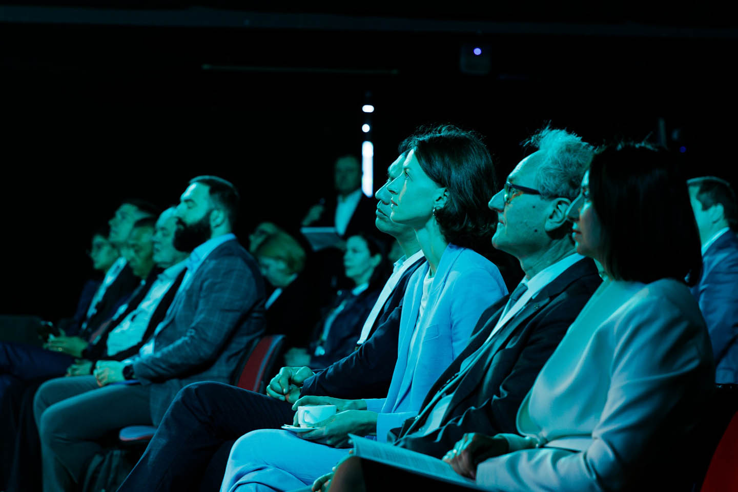 Группа людей в деловой одежде внимательно слушает выступающего на конференции в затемнённом зале.
