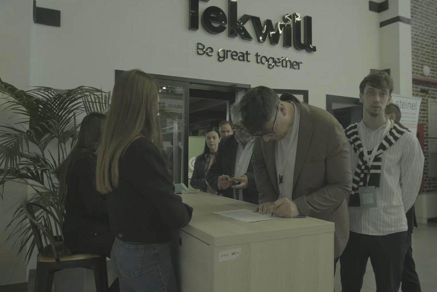 Люди ждут в очереди и регистрируются на стойке в офисе с надписью Tekwill Be great together на стене.