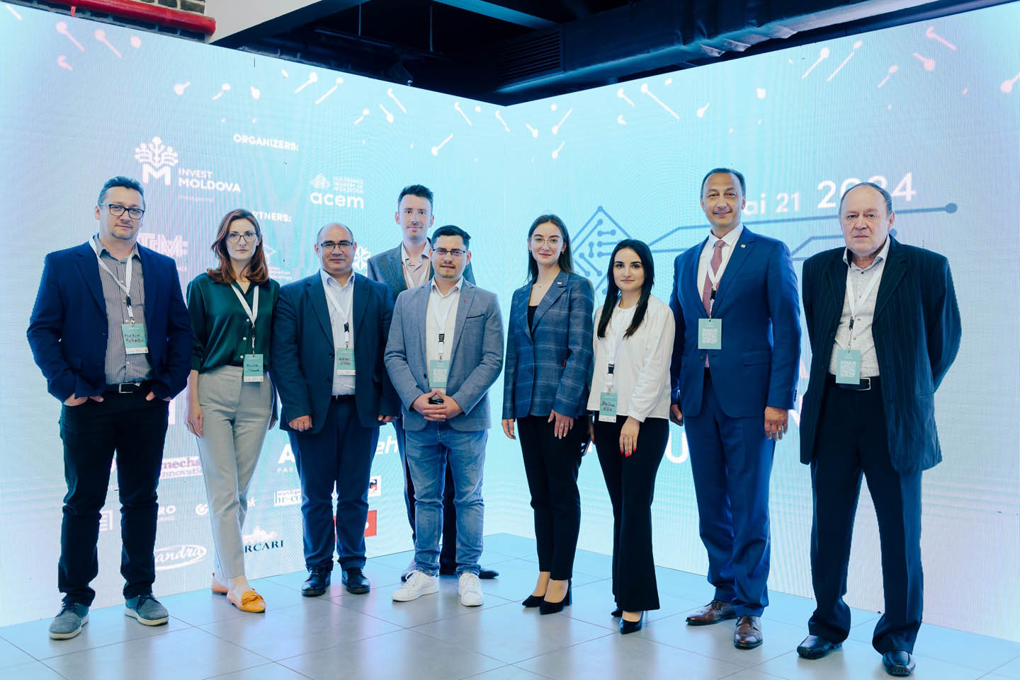 Группа из девяти взрослых, семеро мужчин и две женщины, стоят в ряд на светлом фоне с логотипами и текстом мероприятия.