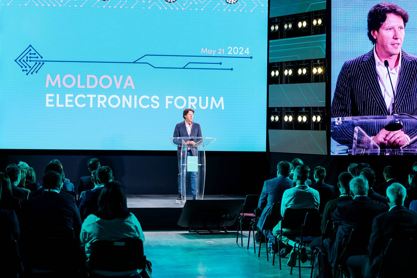 Мужчина в полосатом пиджаке выступает с трибуны на Moldova Electronics Forum перед аудиторией.