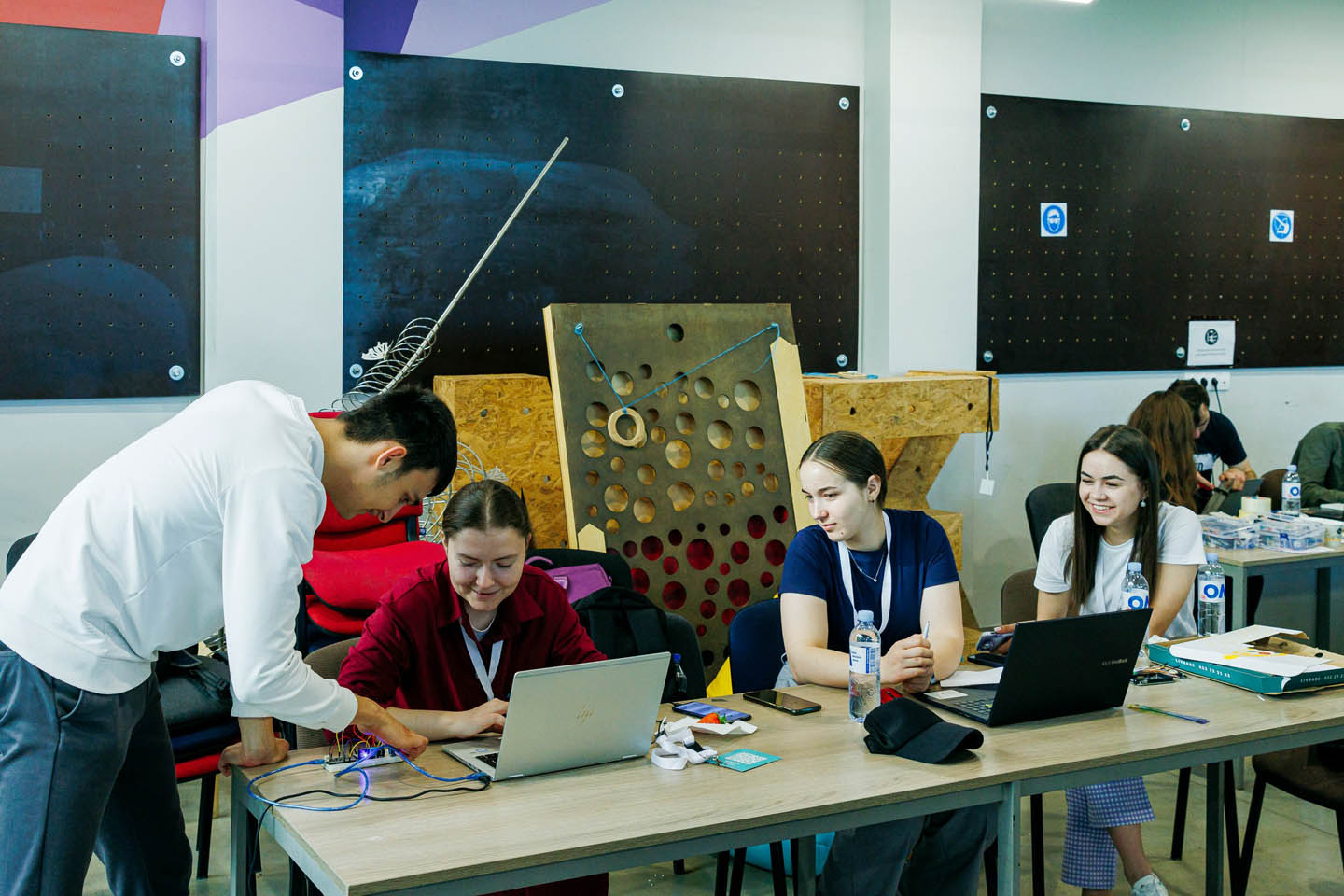 Группа молодых людей работает вместе за столом с ноутбуками и электронными проводами в учебной комнате.