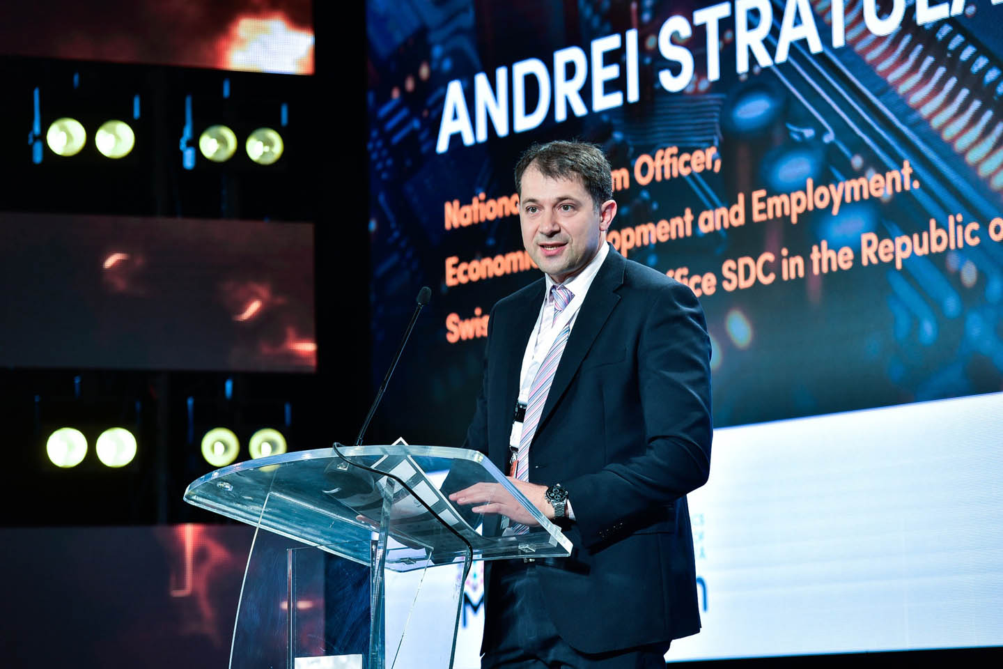 Мужчина в деловом костюме выступает за прозрачной трибуной на конференции с экраном с текстом и световыми приборами на заднем плане.