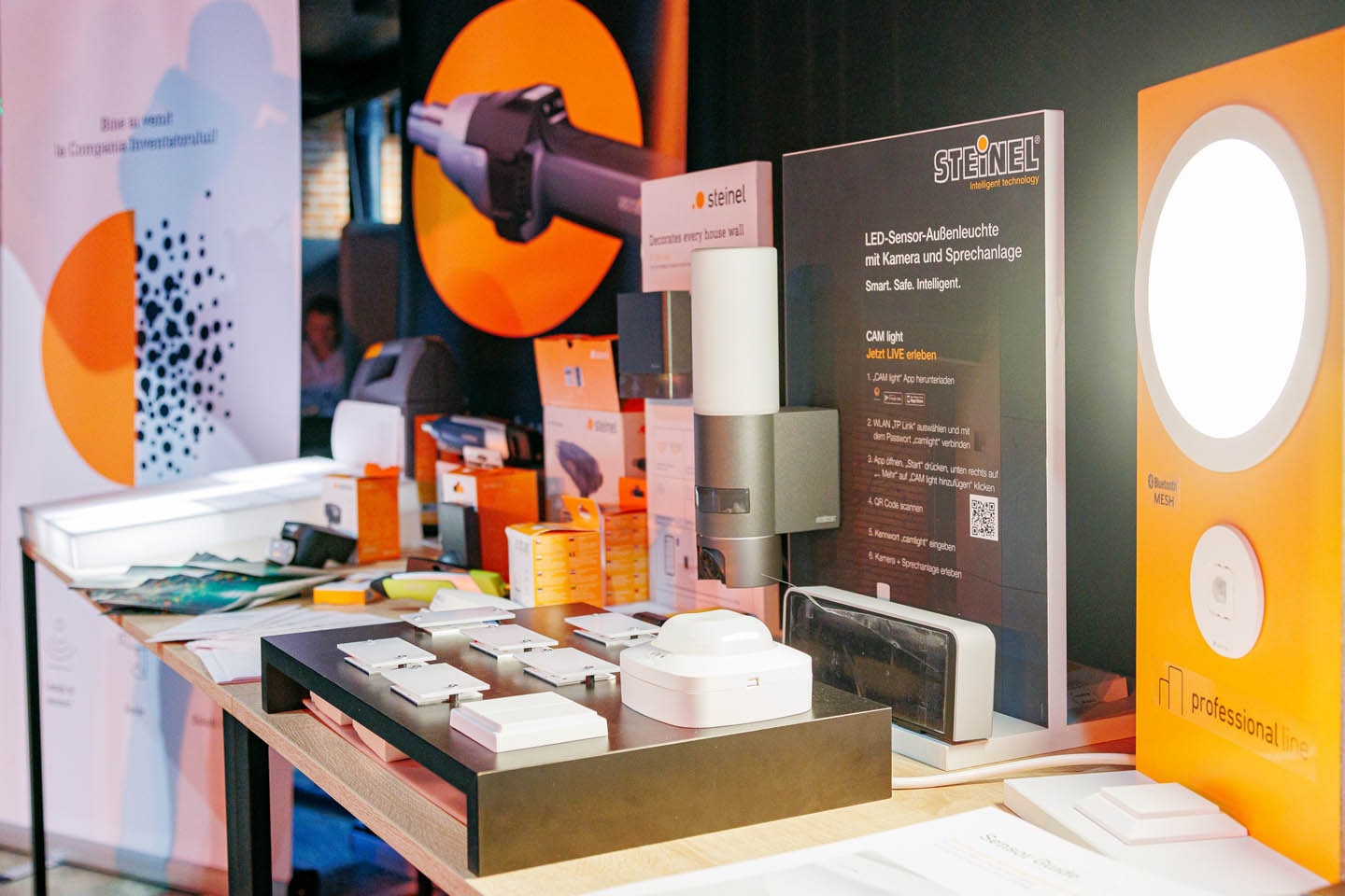 Выставочный стол с различными устройствами и светильниками Steinel, включая светодиодный сенсорный уличный свет с камерой и аудиосистемой.