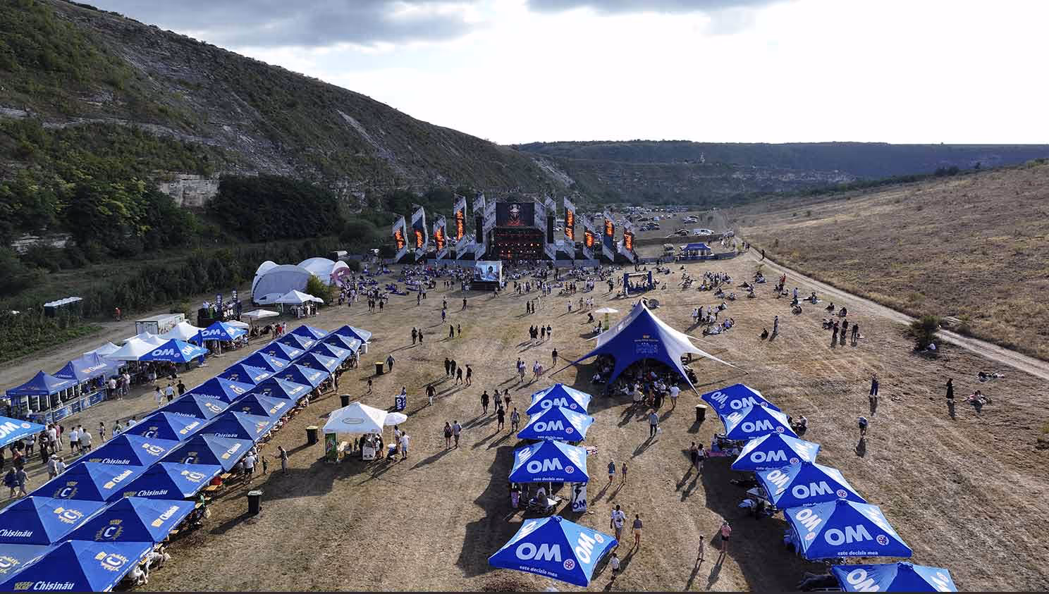 Вид с воздуха на открытый фестиваль с синими палатками и сценой на фоне холмов.