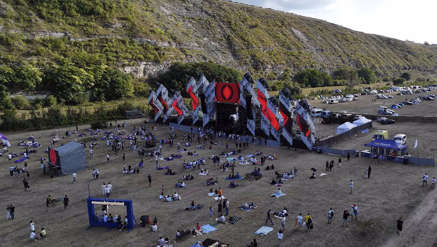 Люди отдыхают на открытом поле перед сценой с яркими геометрическими узорами на фоне зелёного холма.