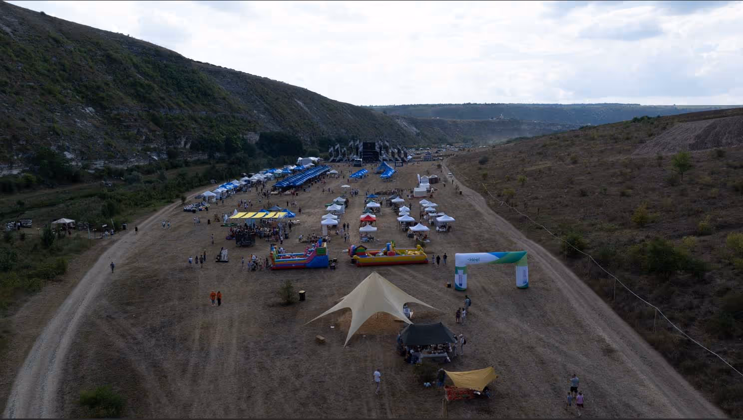 Панорамный вид на фестиваль с палатками, надувными аттракционами и сценой на открытом поле между холмами.