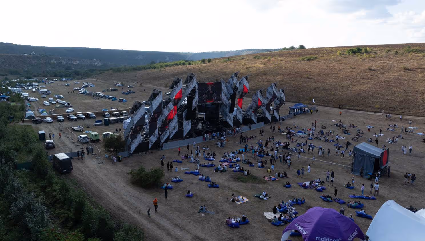 Открытая музыкальная сцена на фестивале на поле у подножия холмов с разбросанными людьми и палатками.