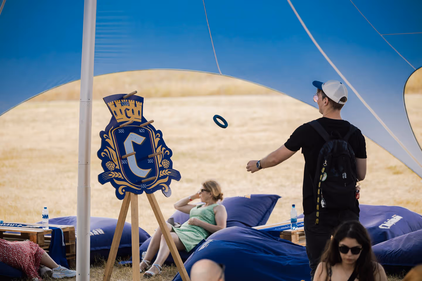 Мужчина в кепке и с рюкзаком играет в метание колец на цель с гербом под шатром, рядом отдыхают женщины на больших синих подушках.