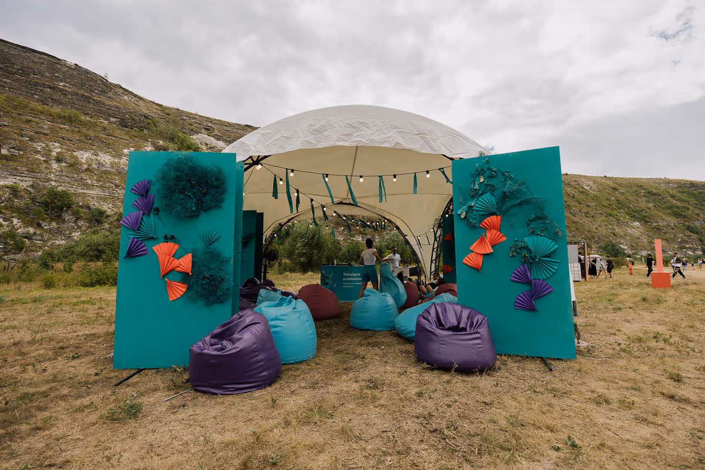 Тентовая конструкция на открытом воздухе с пурпурными и голубыми мешками для сидения на траве, украшенная декоративными вентиляторными элементами, на фоне холмистой местности.