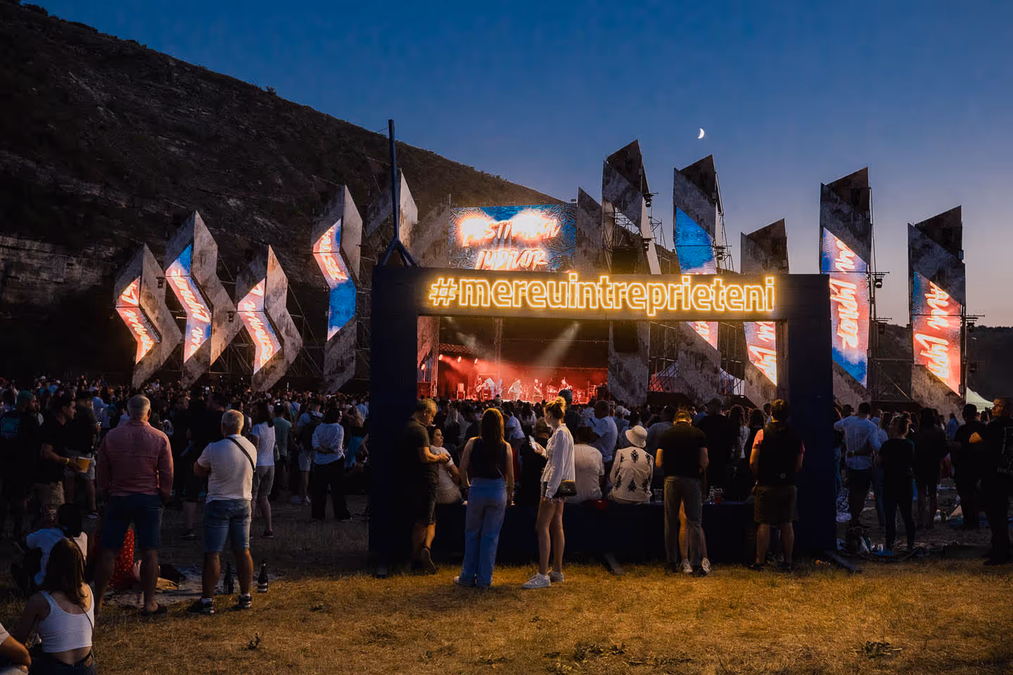 Вечерний музыкальный фестиваль под открытым небом с толпой людей и подсвеченной вывеской #mereuintreprieteni.