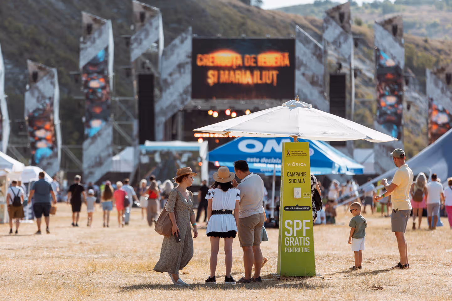 Люди на открытом фестивале, около пункта раздачи бесплатного солнцезащитного крема под зонтом.