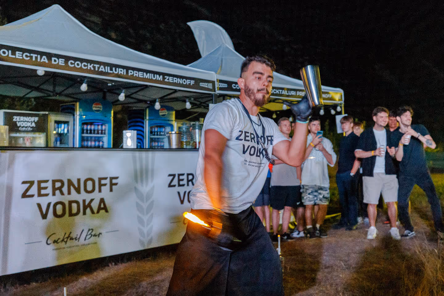 Бармен в футболке Zernoff Vodka жонглирует металлическим шейкером и зажжённым огнём на фоне палатки и группы зрителей.
