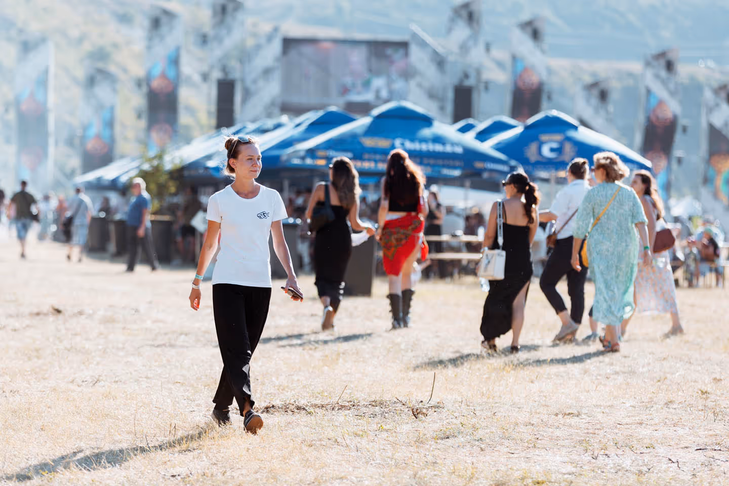 Женщина в белой футболке и черных штанах идет по открытой площадке с другими людьми и синими навесами на заднем плане.
