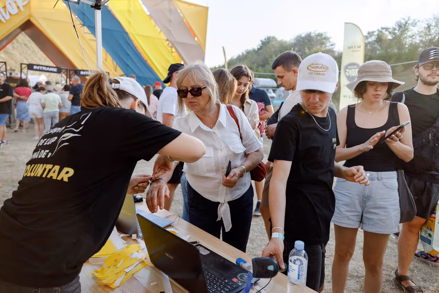 Волонтер надевает браслет на руку пожилой женщины на фестивале, рядом люди стоят в очереди на регистрацию.