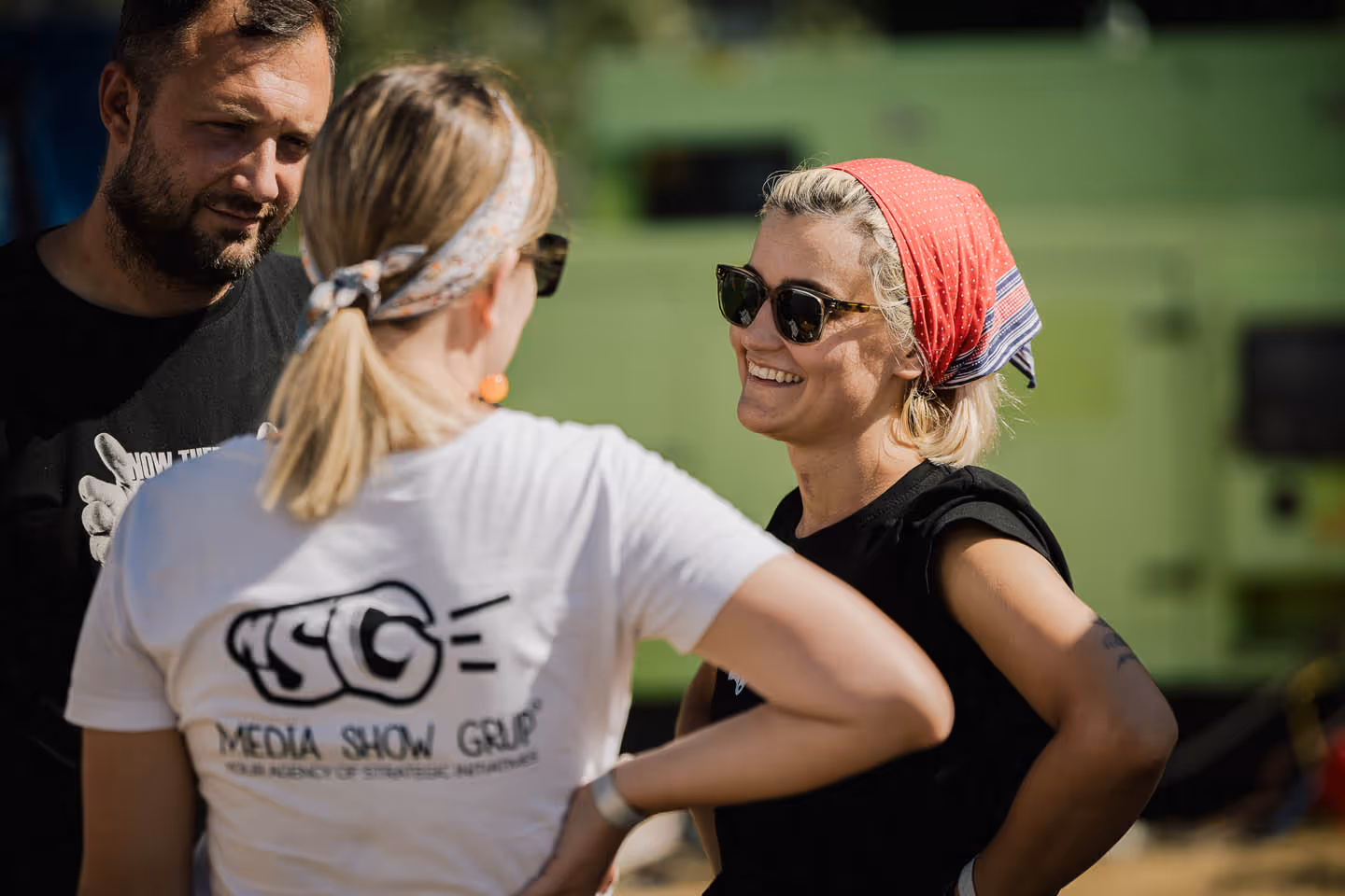 Три человека в солнцезащитных очках ведут дружескую беседу на открытом воздухе, женщина в красной повязке улыбается.