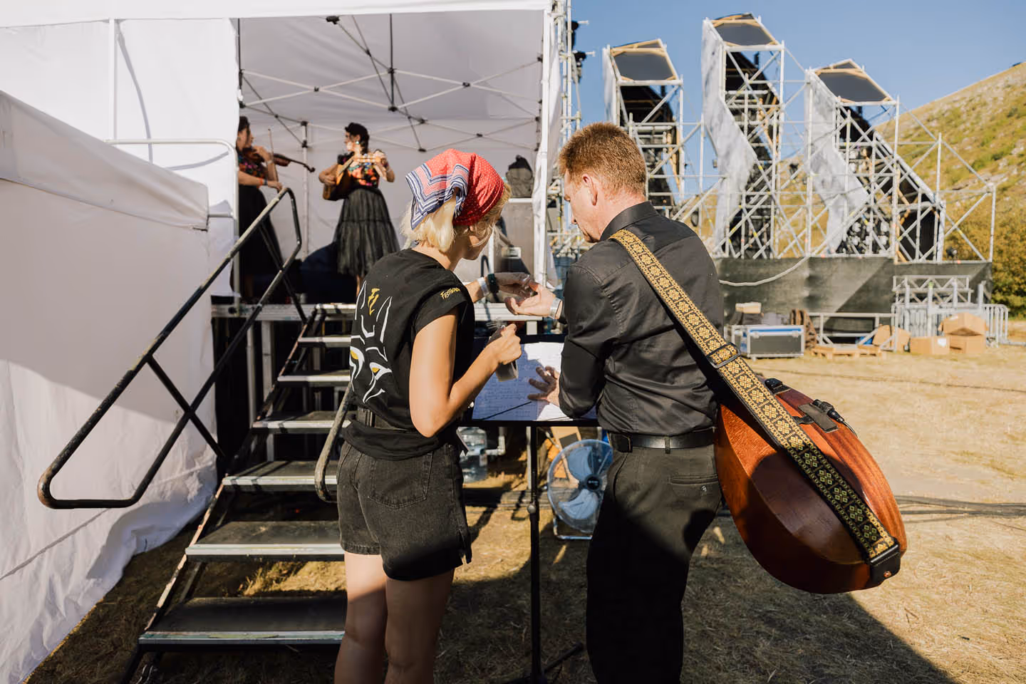 Два музыканта обсуждают ноты у сцены на открытом воздухе, на заднем плане женщины играют на скрипках.