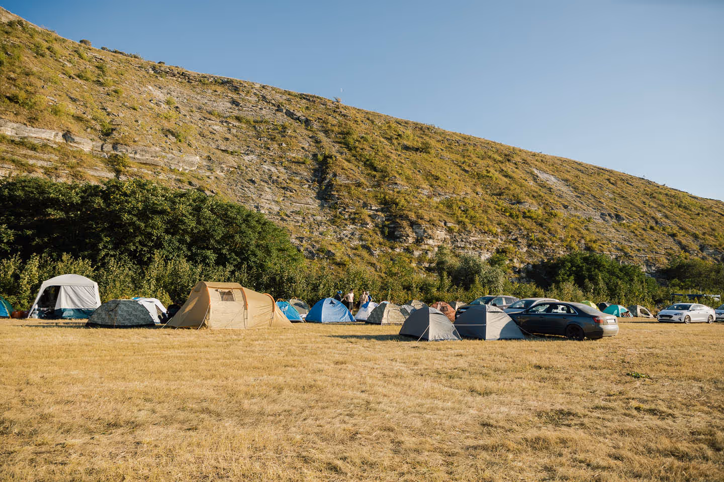 Палаточный лагерь на сухой травяной поляне у подножья холма с несколькими припаркованными автомобилями.