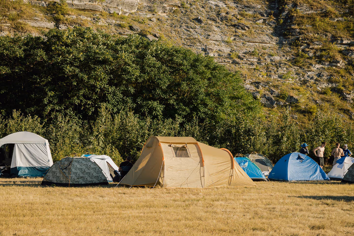 Кемпинг с разными палатками на поляне на фоне холма и густых зеленых деревьев.