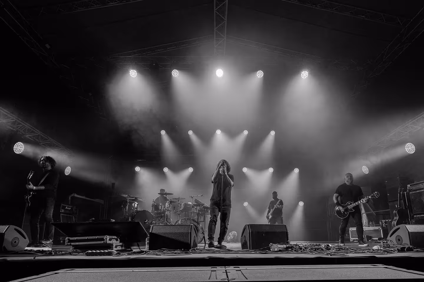 Черно-белое фото музыкальной группы на сцене с вокалистом в центре и гитаристами по бокам под яркими прожекторами.