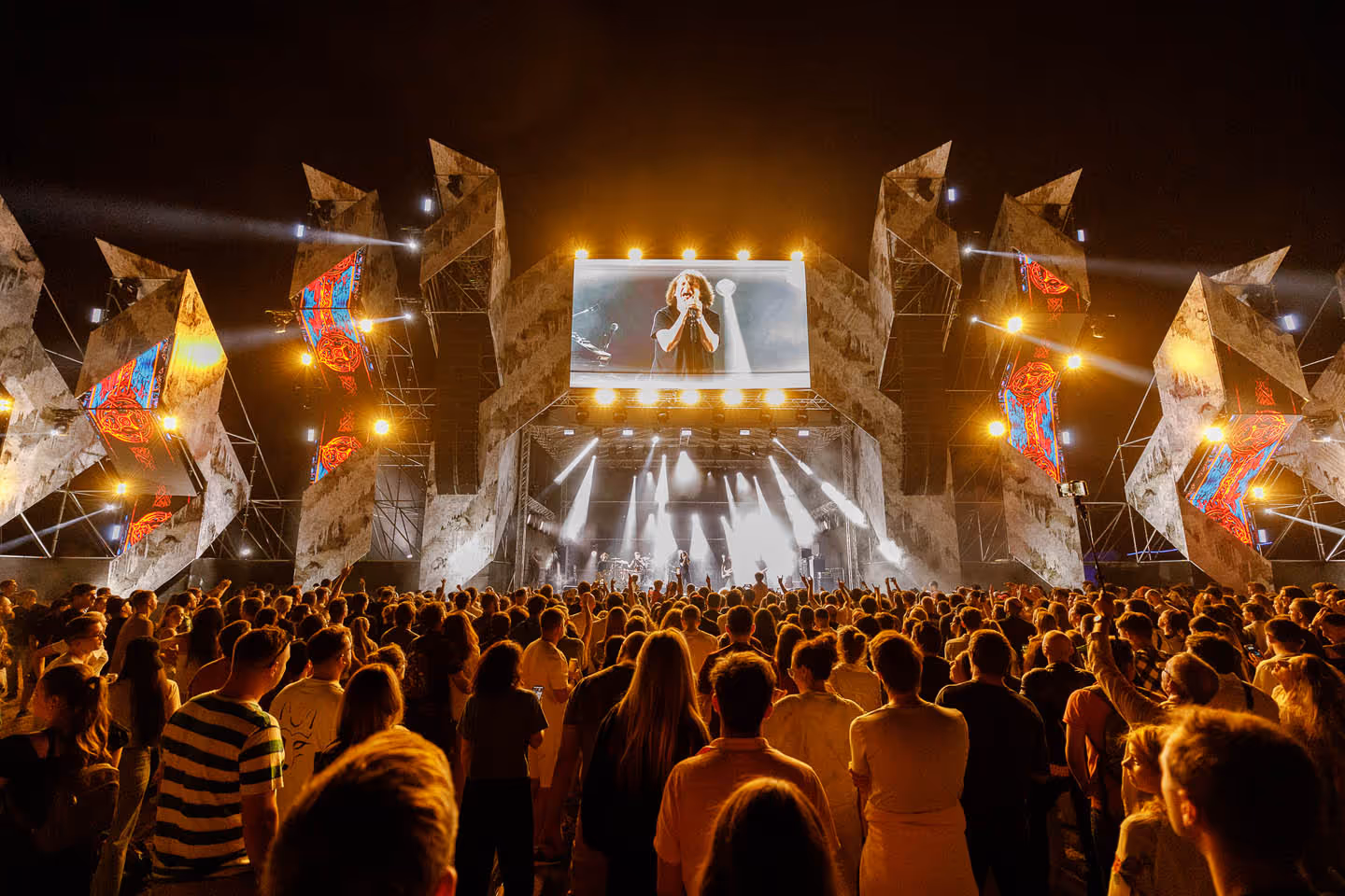 Толпа зрителей на концерте вечером перед освещённой сценой с певцом и яркими прожекторами.