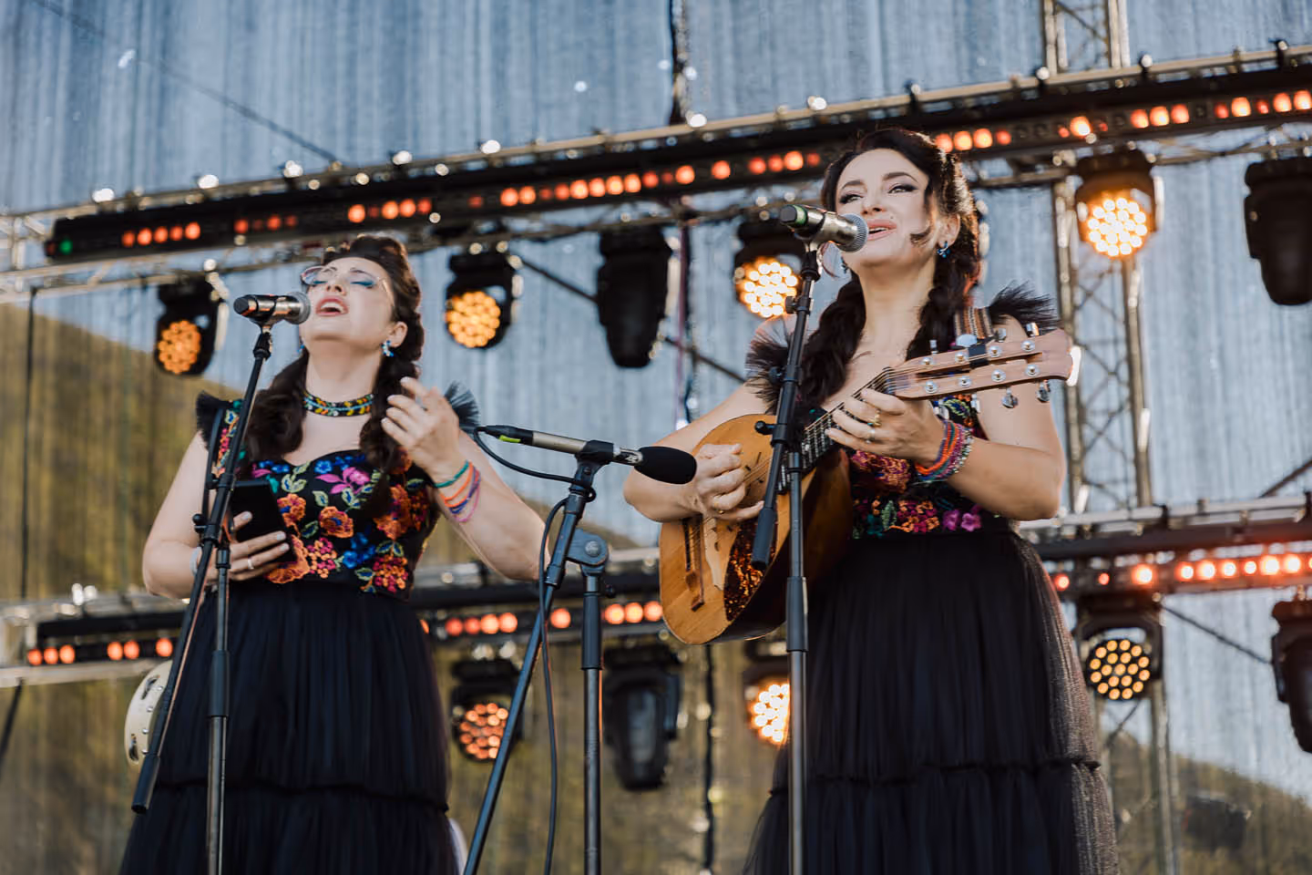 Две женщины в черных платьях с ярким цветочным узором поют на сцене, одна из них играет на гитаре.