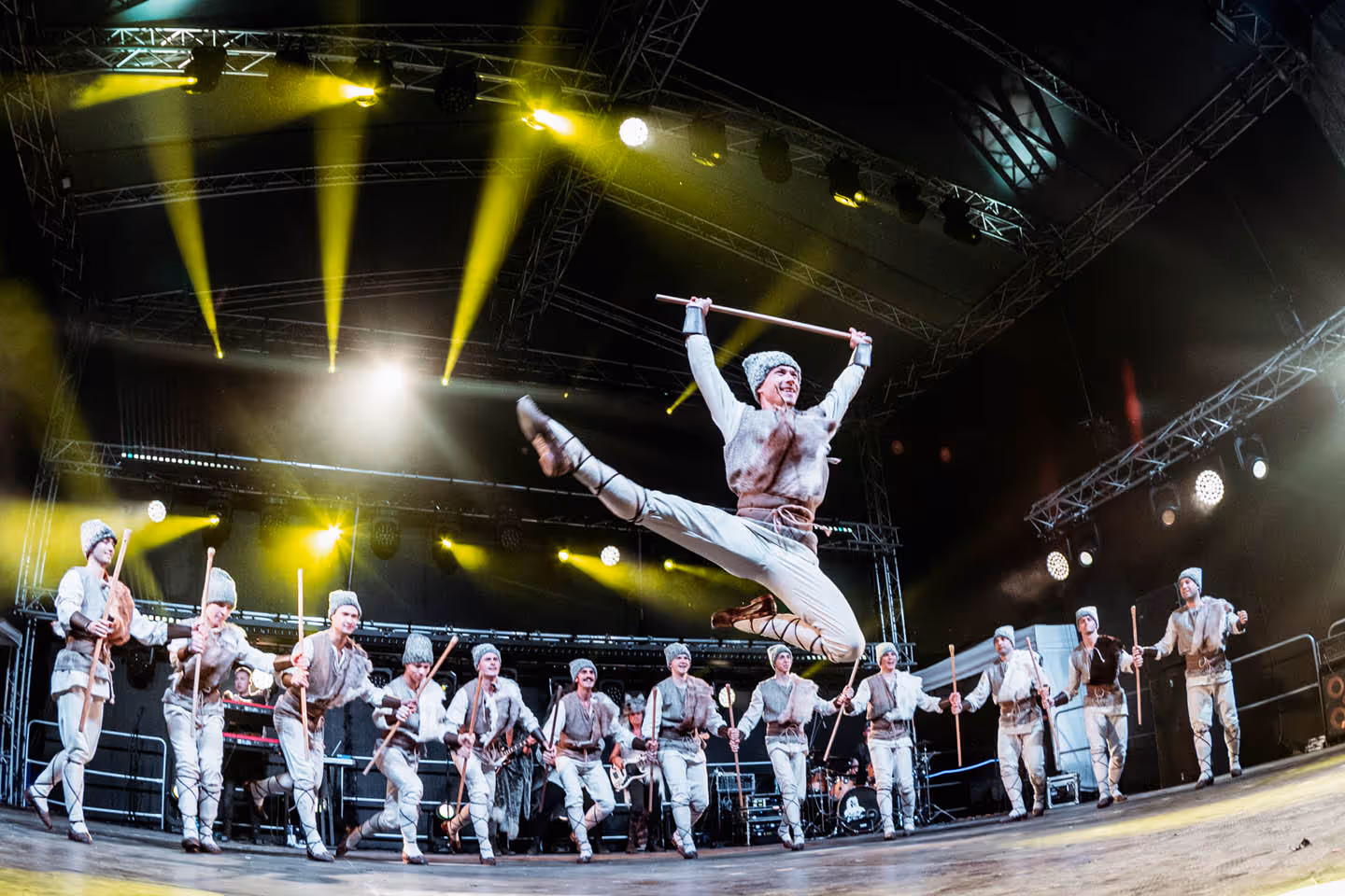 Танцоры в национальной одежде с палками выступают на сцене под жёлтым сценическим светом, один из них прыгает в прыжке с широко разведёнными ногами.