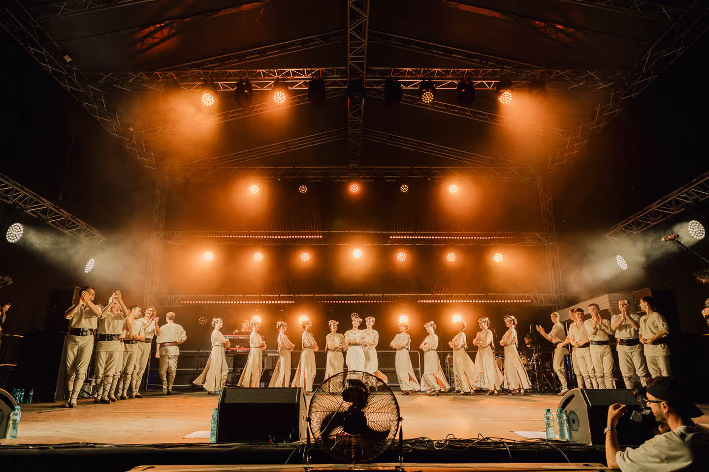 Группа танцоров в светлых костюмах и венках на сцене под оранжевыми прожекторами.