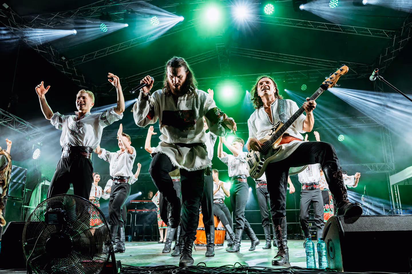 Группа музыкантов и танцоров в традиционной одежде выступает на сцене с ярким зелёным освещением.