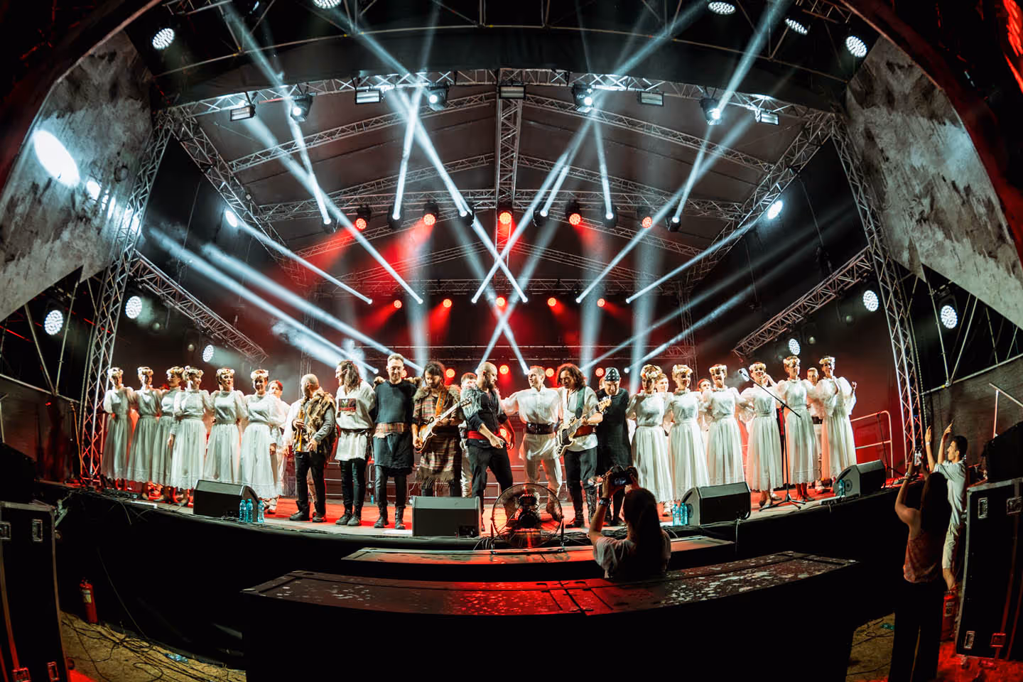 Группа из музыкантов и певцов в традиционных костюмах на сцене с белыми и красными прожекторами.