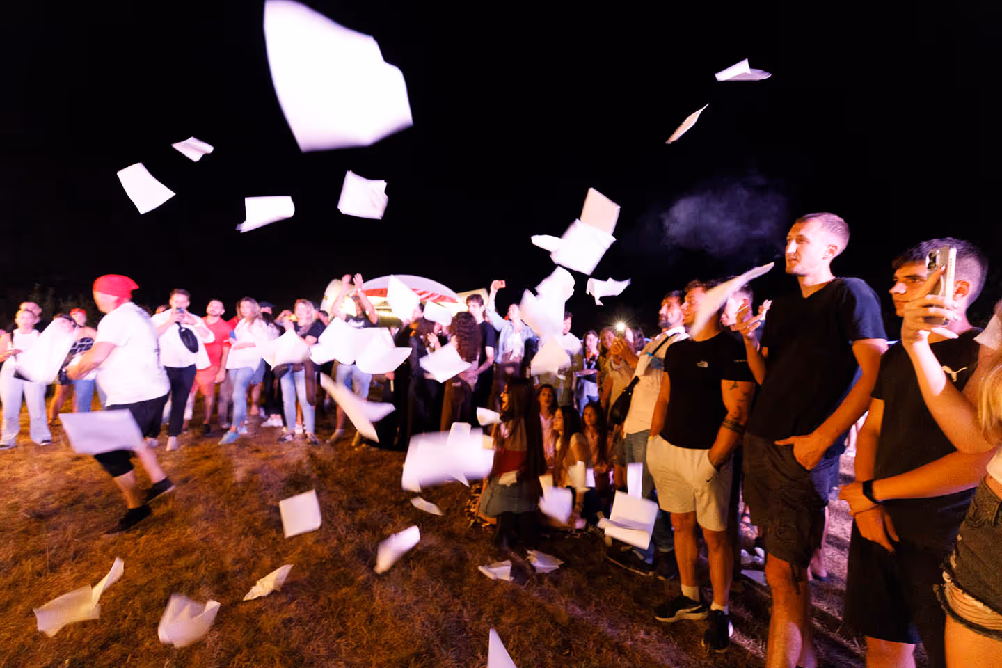 Толпа людей на ночном открытом мероприятии, вокруг летают воздушные белые листы бумаги.