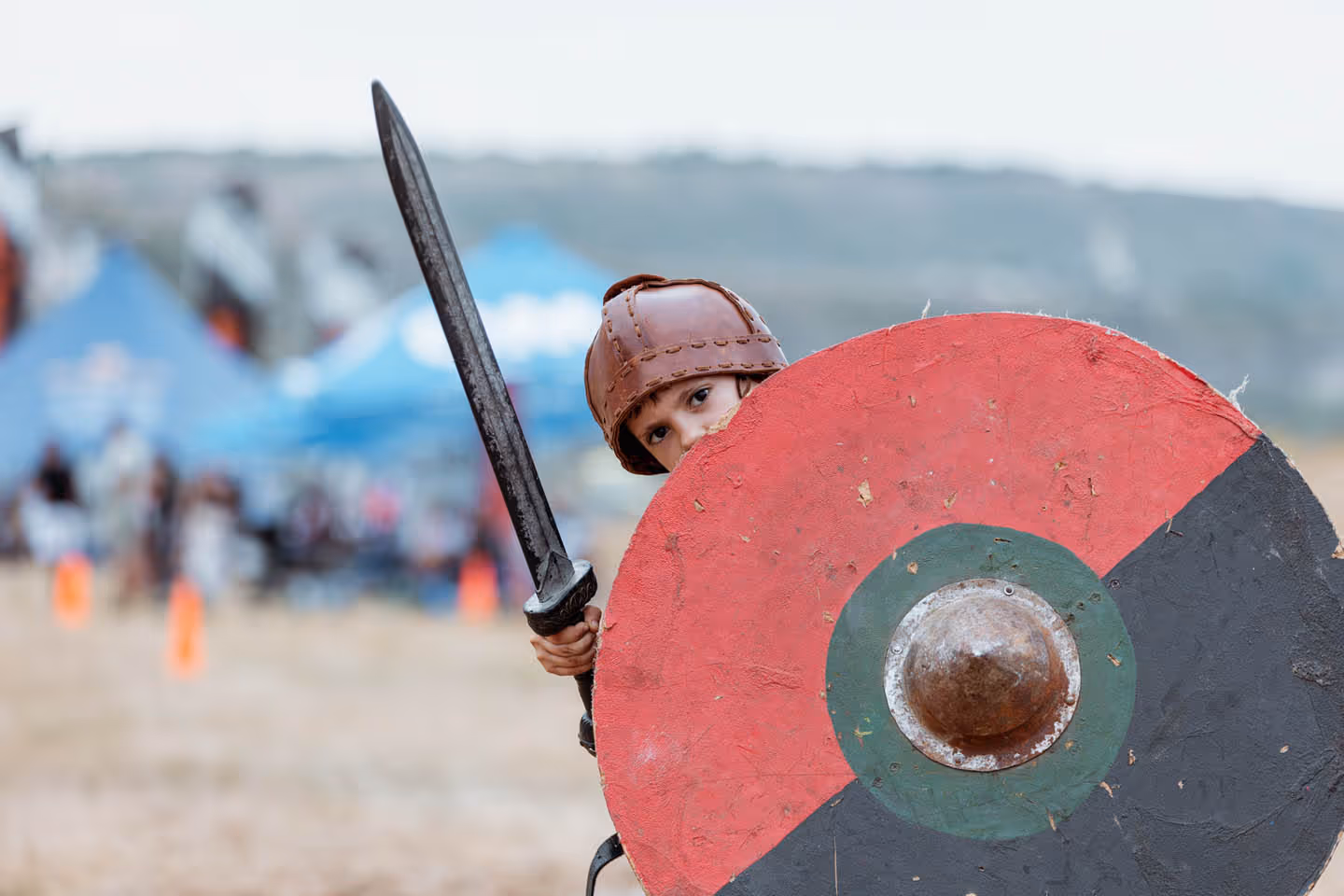 Ребёнок в коричневом шлеме прячется за круглый щит с красной и чёрной окраской, держа в руках меч.