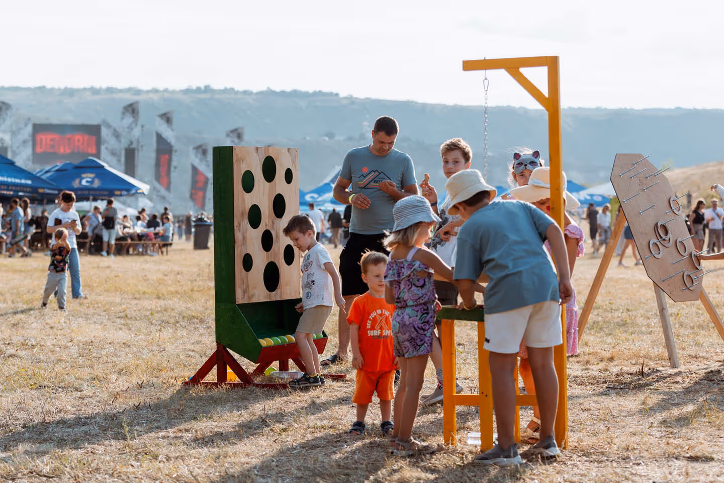 Группа детей и взрослый играют на открытом воздухе с большими деревянными играми на травянистом поле во время фестиваля.