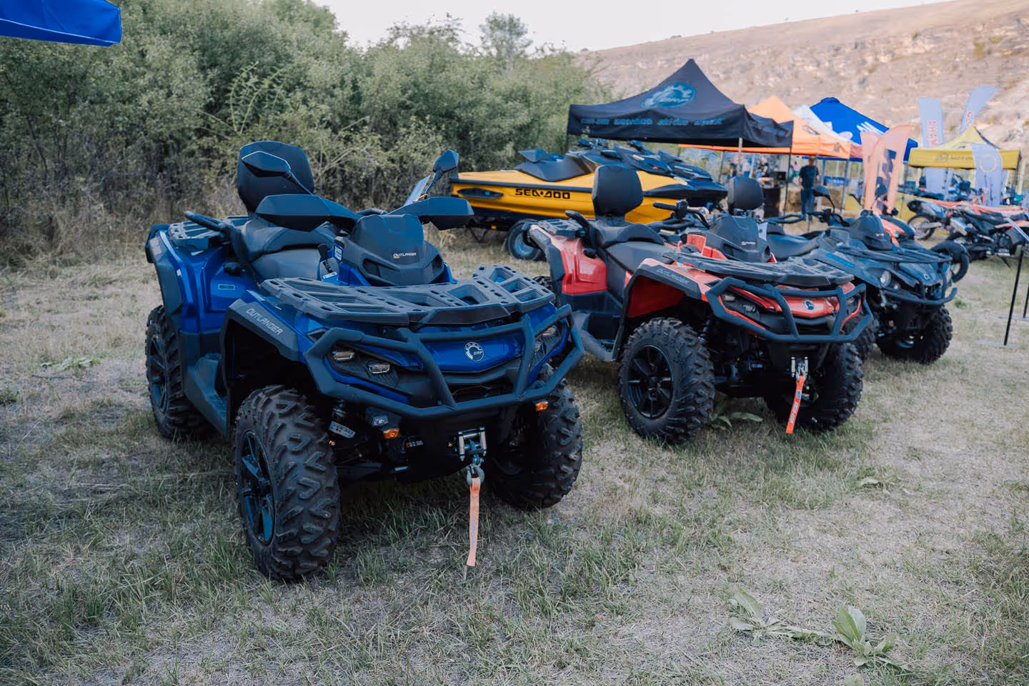 Ряды синих и красных квадроциклов, выставленных на открытом воздухе на травянистой площадке с палатками и людьми на заднем плане.