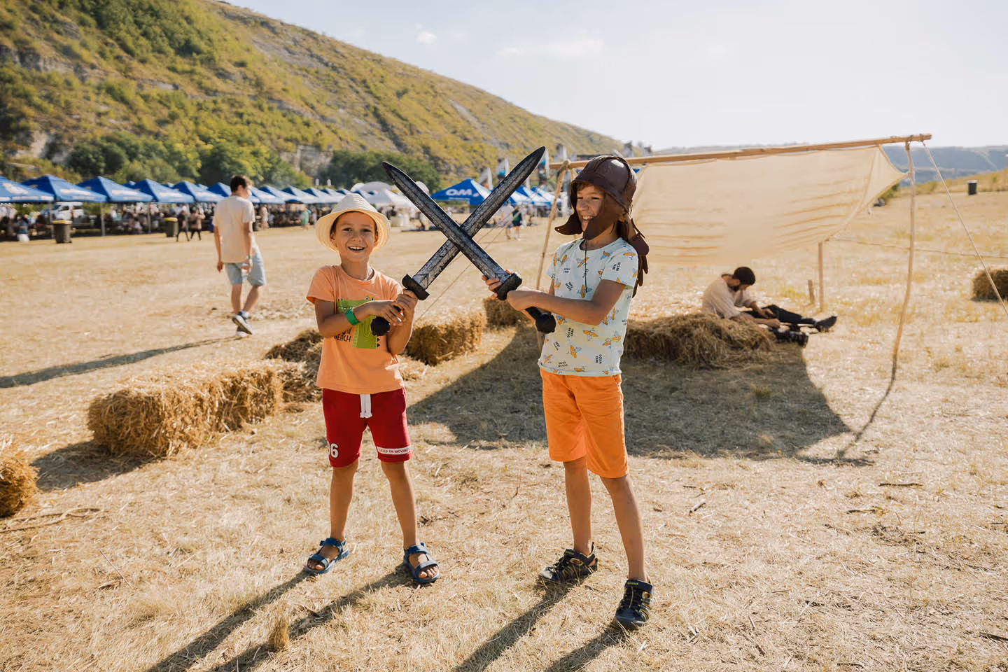 Два ребенка играют с пластиковыми мечами на открытом воздухе, один в шляпе, другой в кожаной шапке, на фоне сена и шатров.