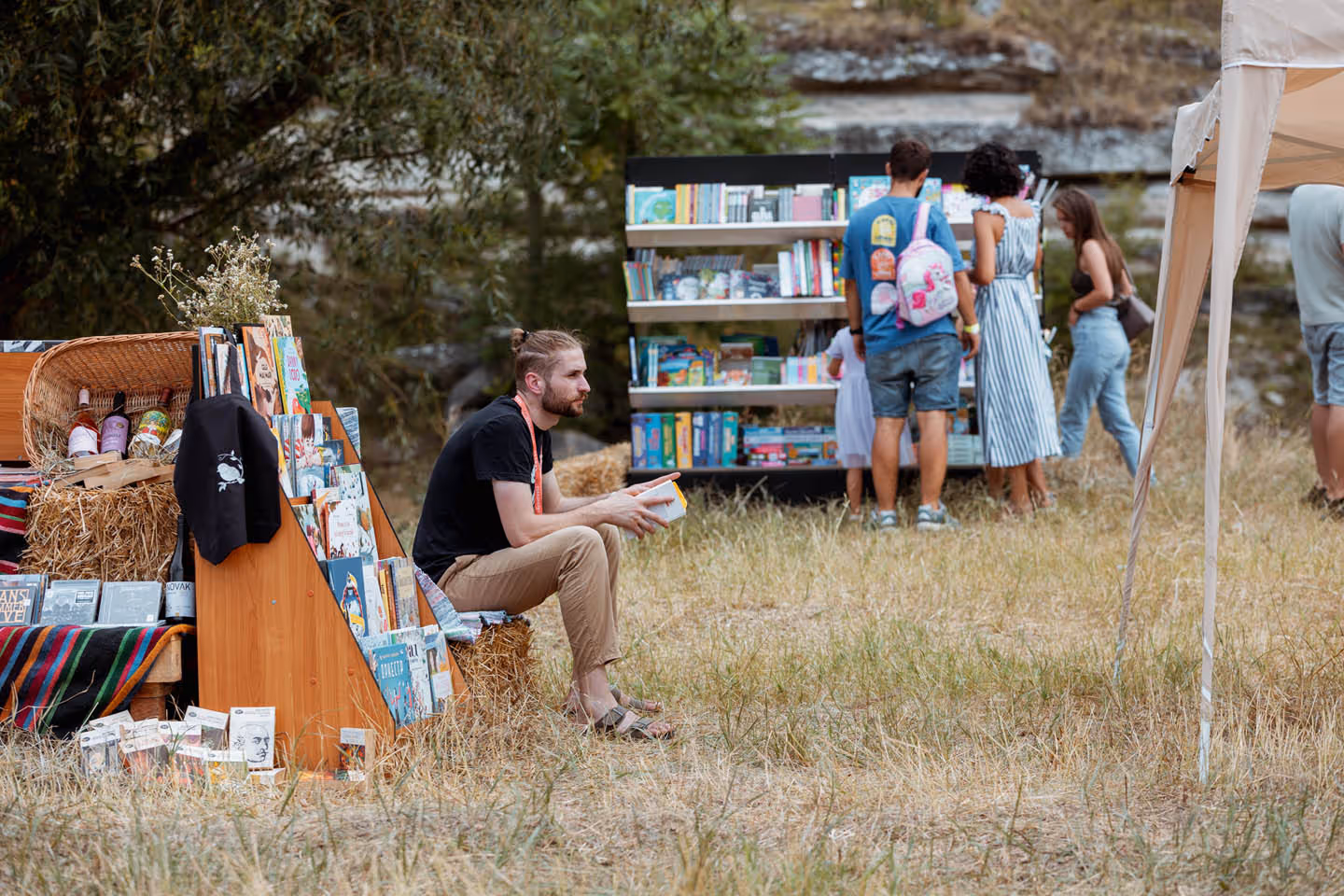 Мужчина сидит на сене, читая книгу у стенда с книгами, на заднем плане люди выбирают книги на открытом воздухе.