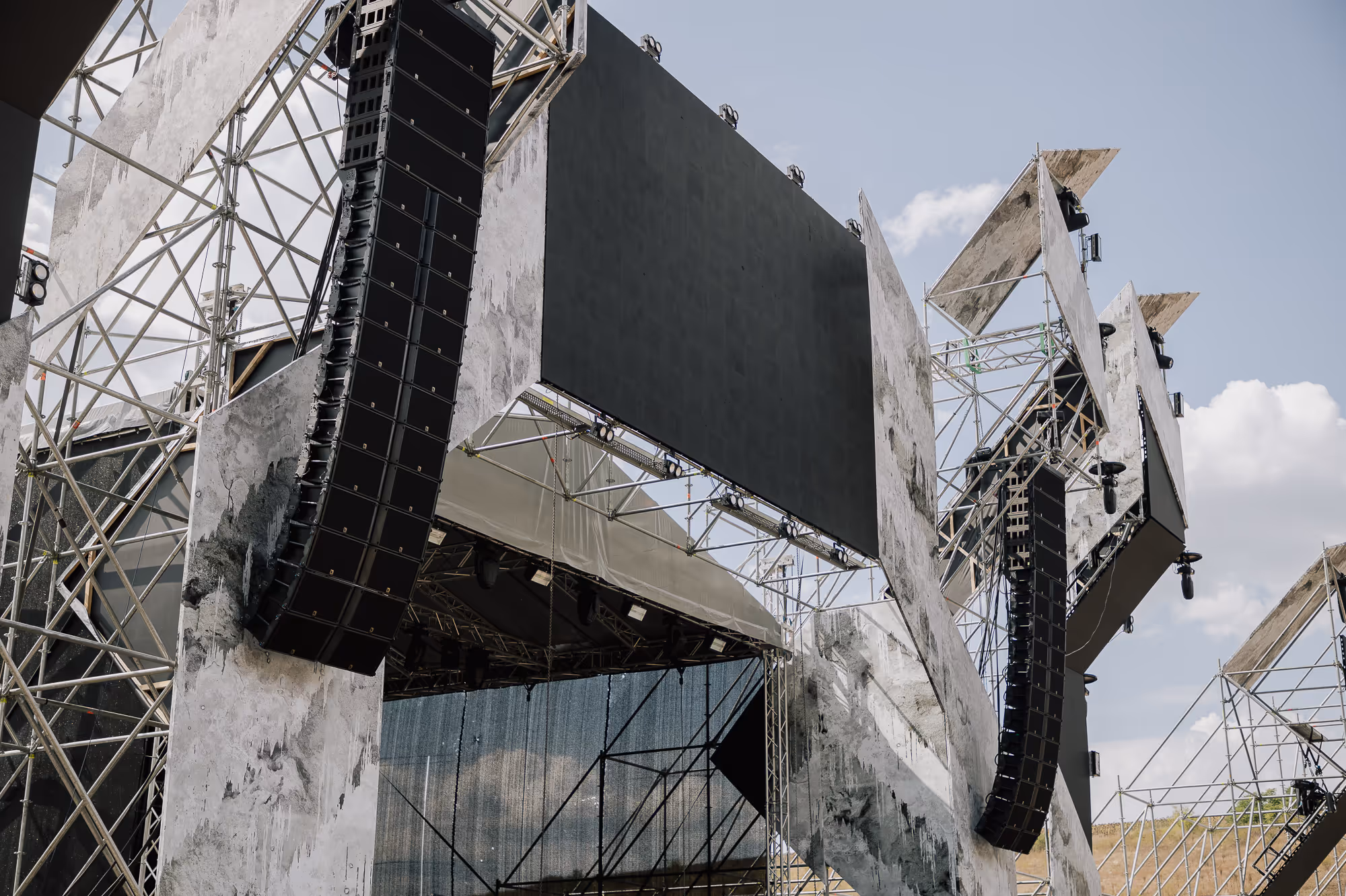 Большая уличная сцена с массивными черными динамиками, крупным экраном и металлическими конструкциями на фоне голубого неба с облаками.