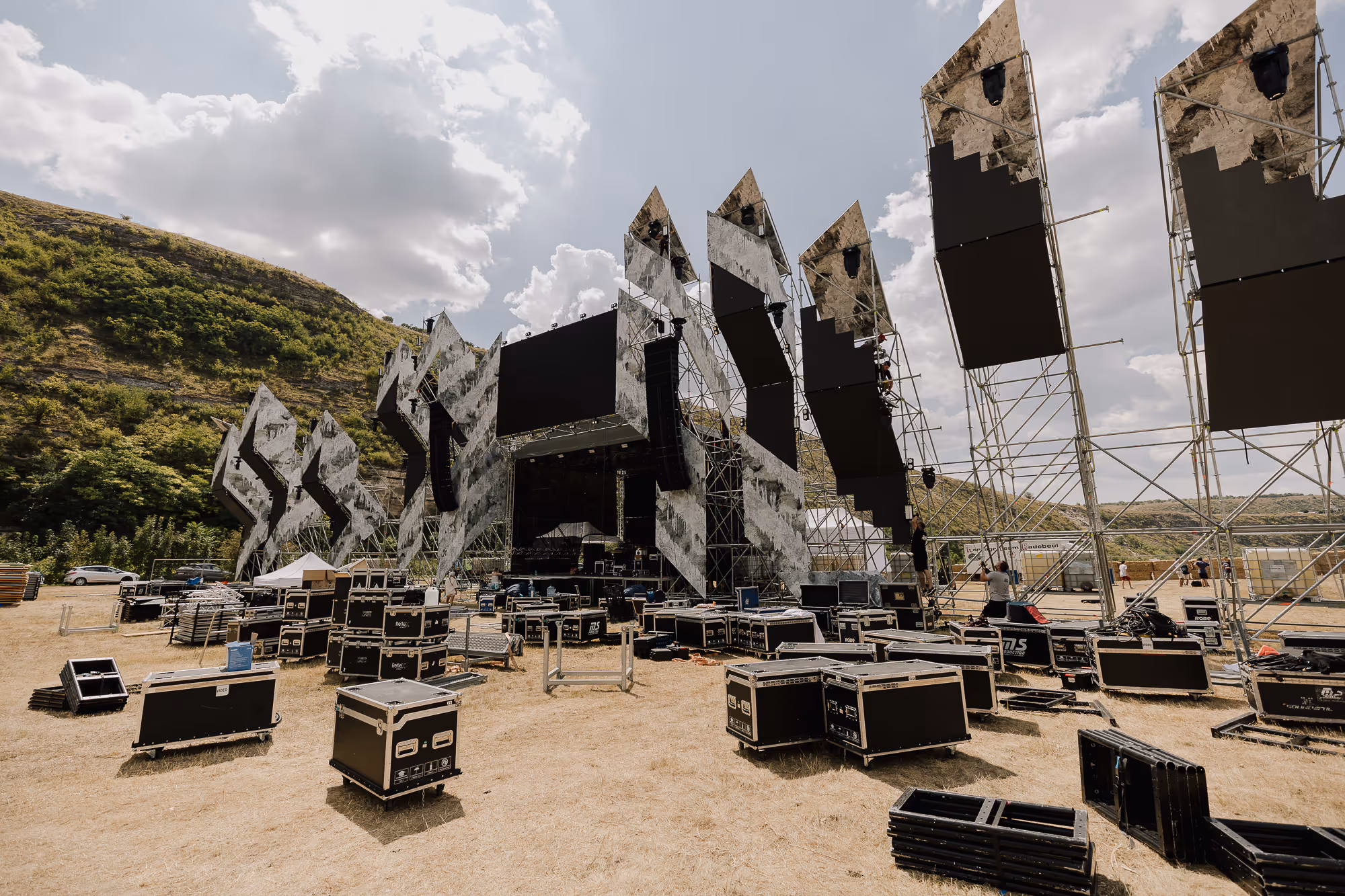 Большая сцена на открытом воздухе с черными экранными панелями и готовящимся оборудованием перед музыкальным фестивалем в холмистой местности.