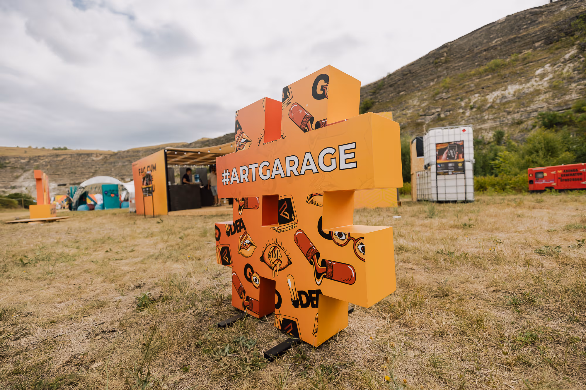 Оранжевая скульптура в форме хэштега с надписью #ARTGARAGE и рисунками красных губ, глаз и ламп, установленная на травянистом поле с горой на заднем плане.