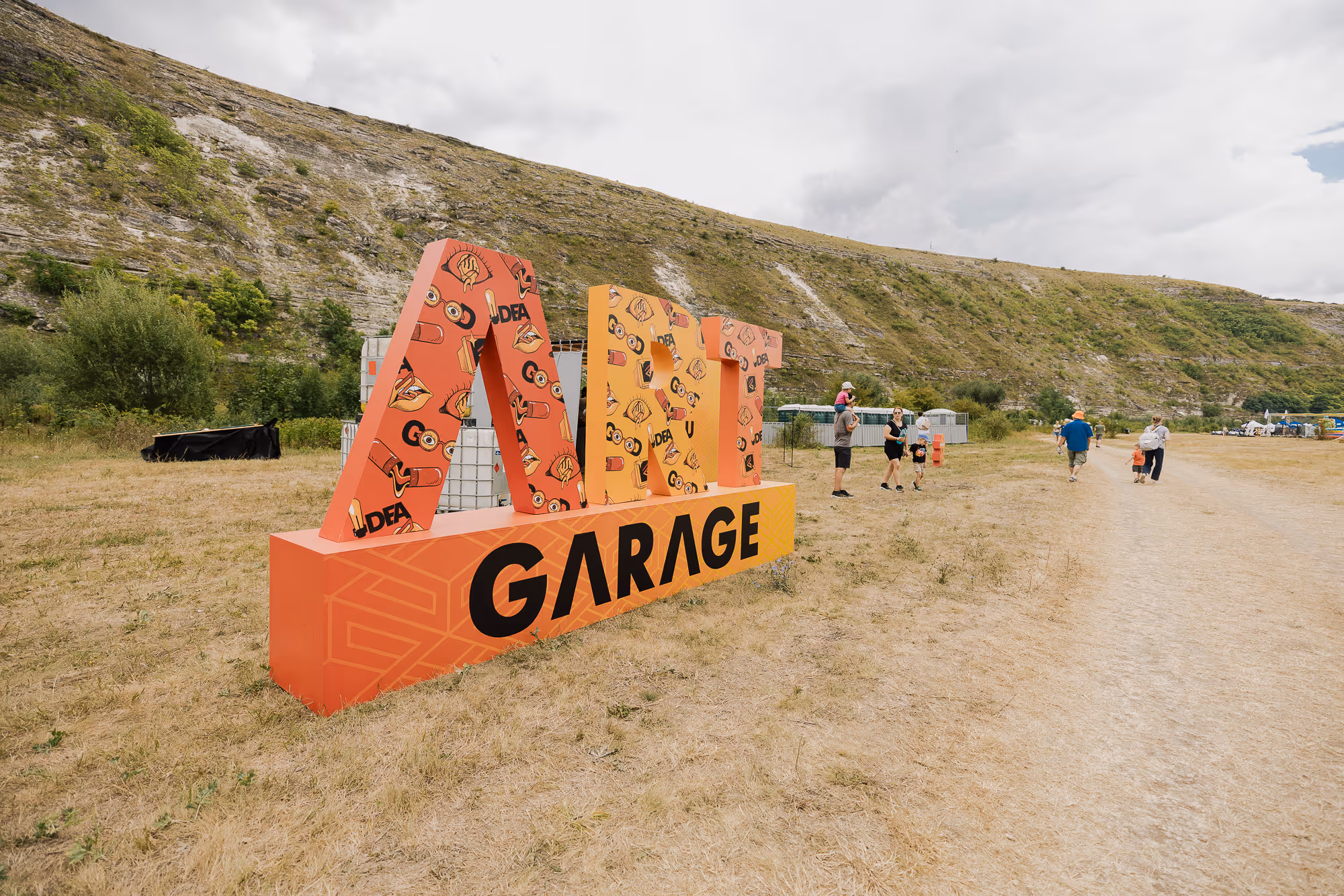 Яркая большая скульптура с надписью ART GARAGE на фоне холма и людей на открытом воздухе.