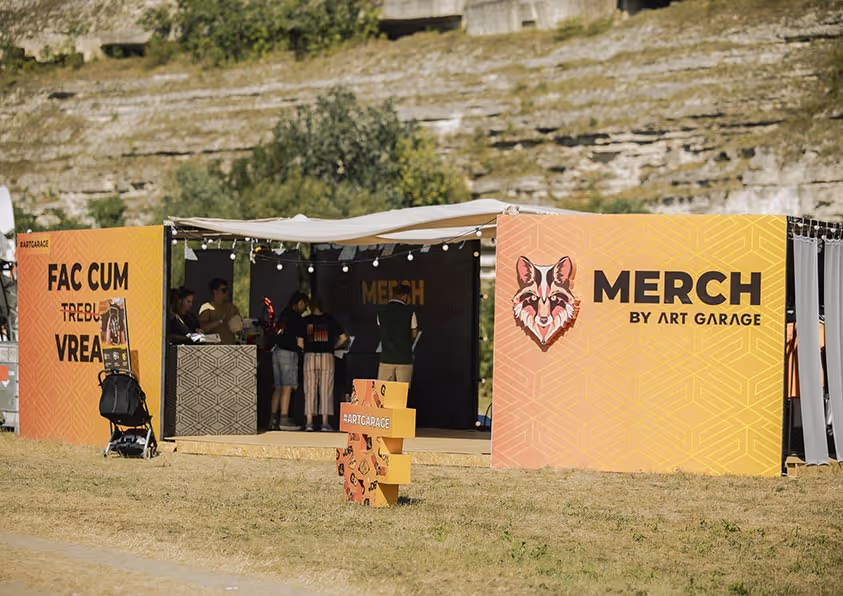 Открытая палатка с оранжевыми стендами для продажи мерча Art Garage на фестивале, несколько человек стоят внутри.