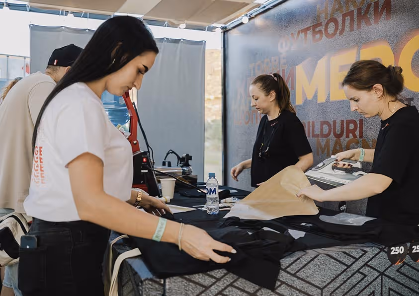 Четыре человека работают с черными футболками на столе под крышей, одна женщина гладит футболку утюгом.