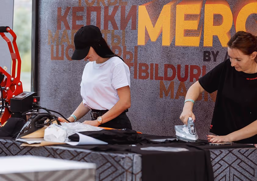 Две женщины гладят одежду на столах с черной тканью на фоне стены с надписью Merc.