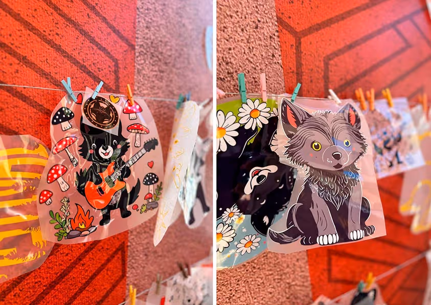 Рисунки в прозрачных конвертах с изображениями мультяшных волков висят на бельевой верёвке с прищепками на фоне красной стены.