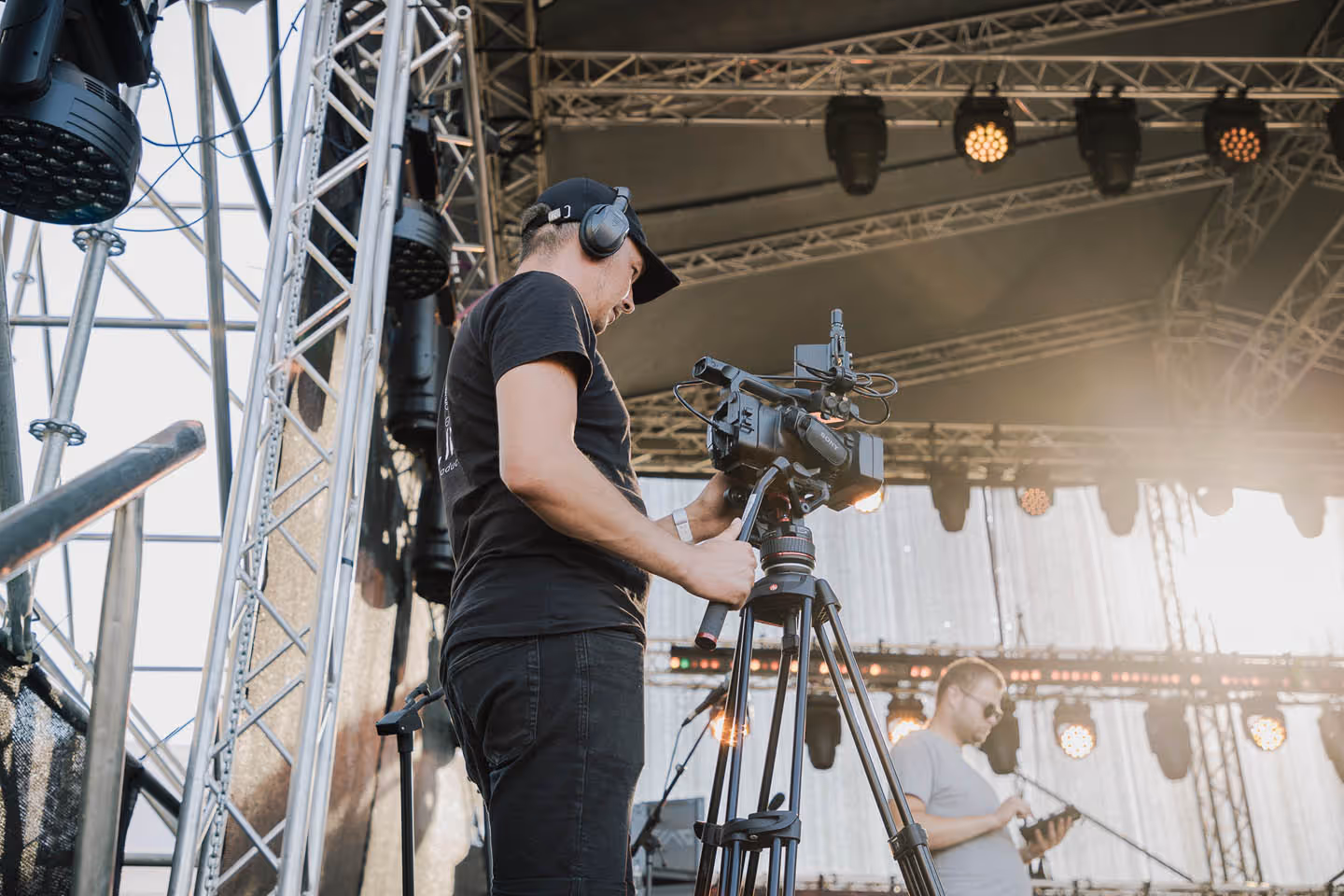 Оператор с наушниками снимает видео на профессиональную камеру на штативе на сцене под навесом.