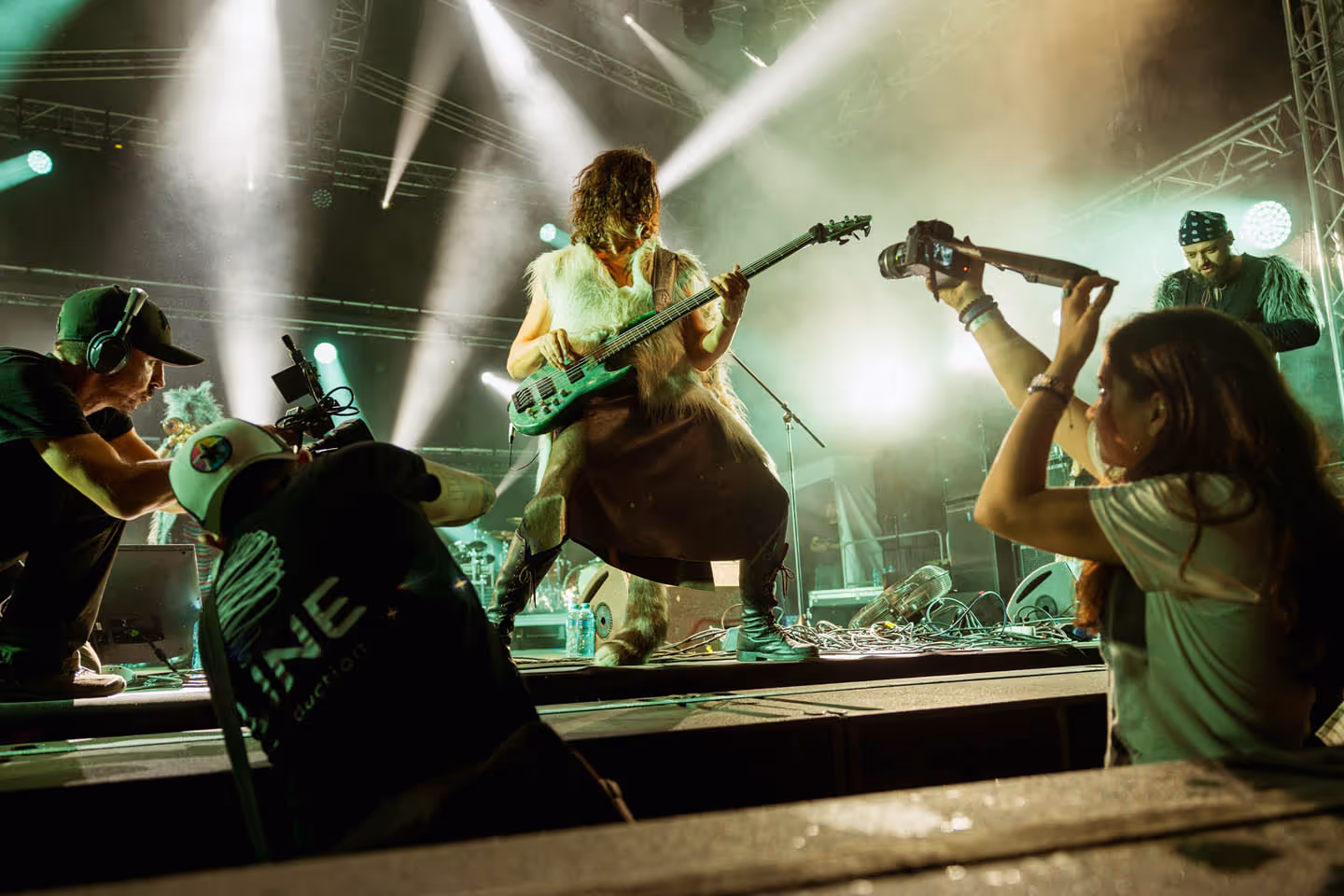 Музыкант в меховом жилете играет на бас-гитаре на сцене во время концерта с освещением и операторами с камерами.