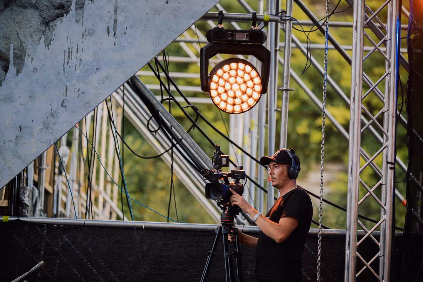 Оператор в черной футболке и кепке с наушниками управляет видеокамерой на штативе под сценическим светом.