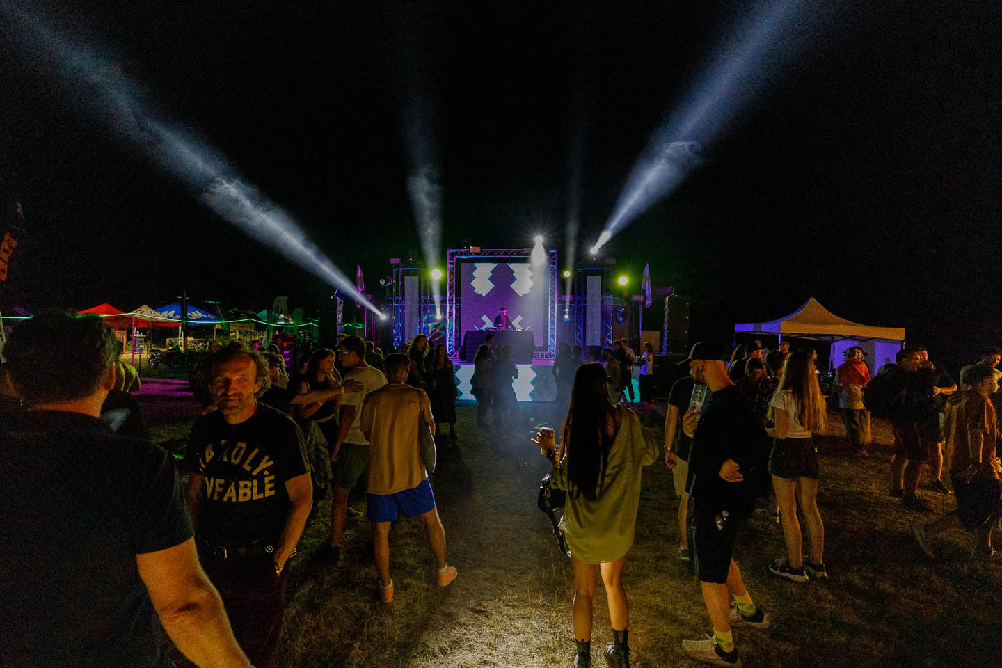 Ночная сценa с ди-джеем и разноцветными прожекторами, люди танцуют и общаются на открытом воздухе.