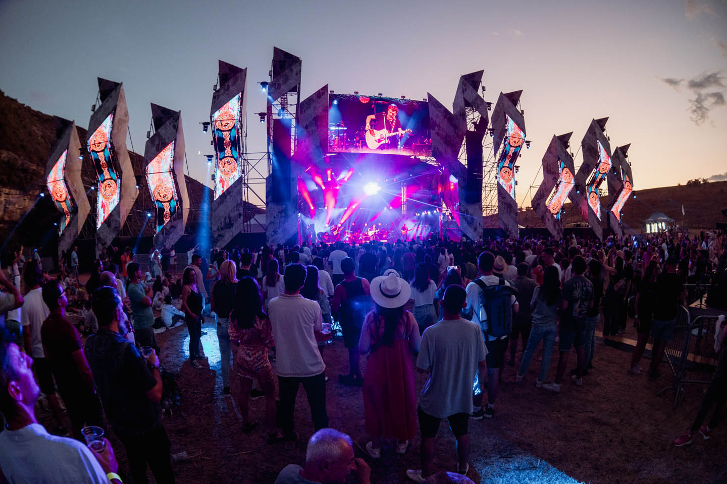 Большая толпа людей смотрит на вечерний концерт с подсветкой и экраном, показывающим исполнителя с гитарой.